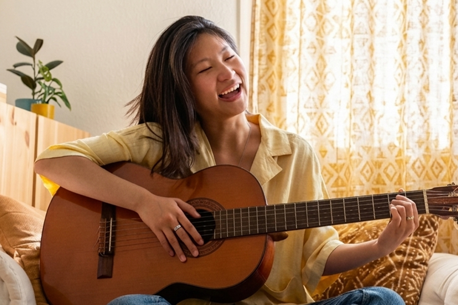 Woman playing an acoustic guitar and singing joyfully while sitting on a couch at home, with warm sunlight coming through the window behind her.