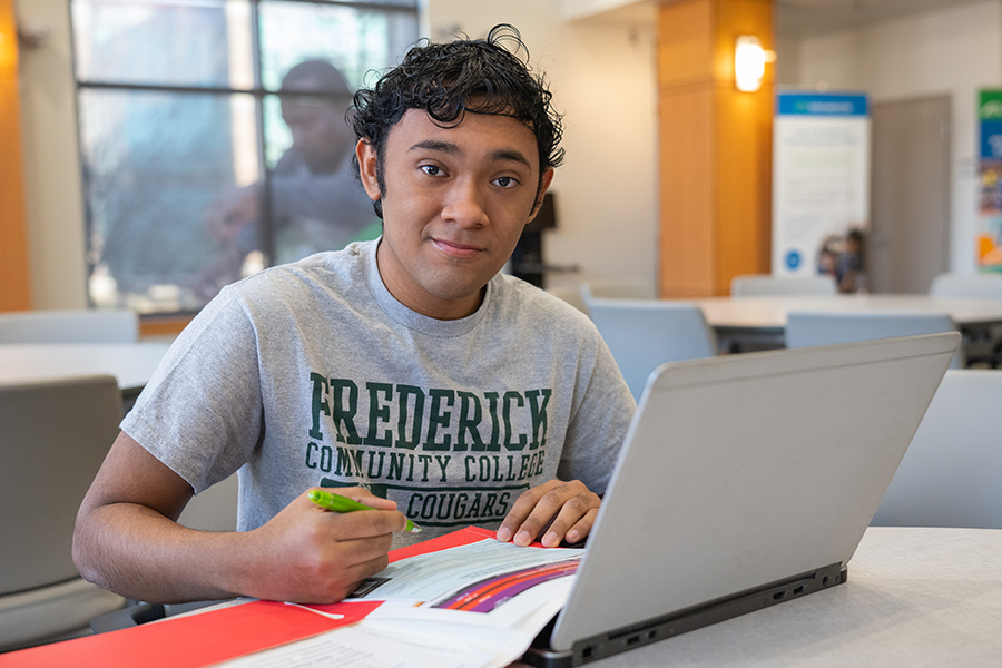 A smiling male student wearing a grey Frederick Community College t-shirt sits at a table with a laptop and a red folder, holding a pen as if ready to work.
