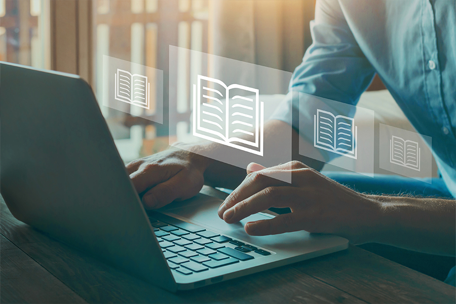A person’s hands typing on a laptop on a wooden table. Semi-transparent digital icons of open books float above the keyboard, representing a digital search or online catalog interface.